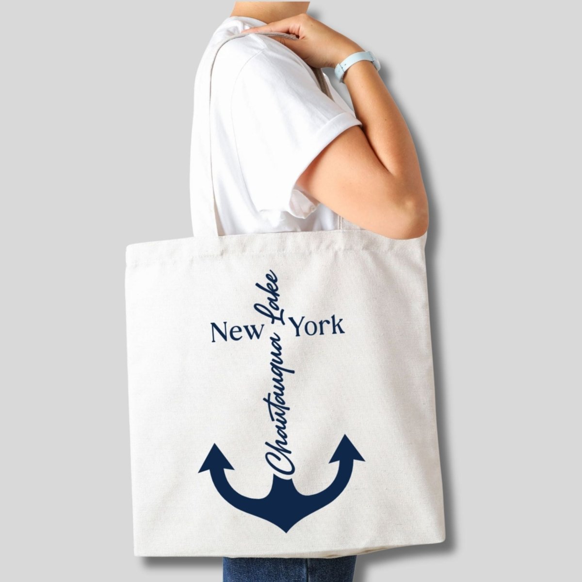 Person holding a creme tote bag with navy anchor design "Chautauqua Lake"on front on gray background - Little Gift Nook