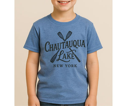 Child wearing blue tshirt with Chautauqua Lake New York and paddles on front standing on tan white background.