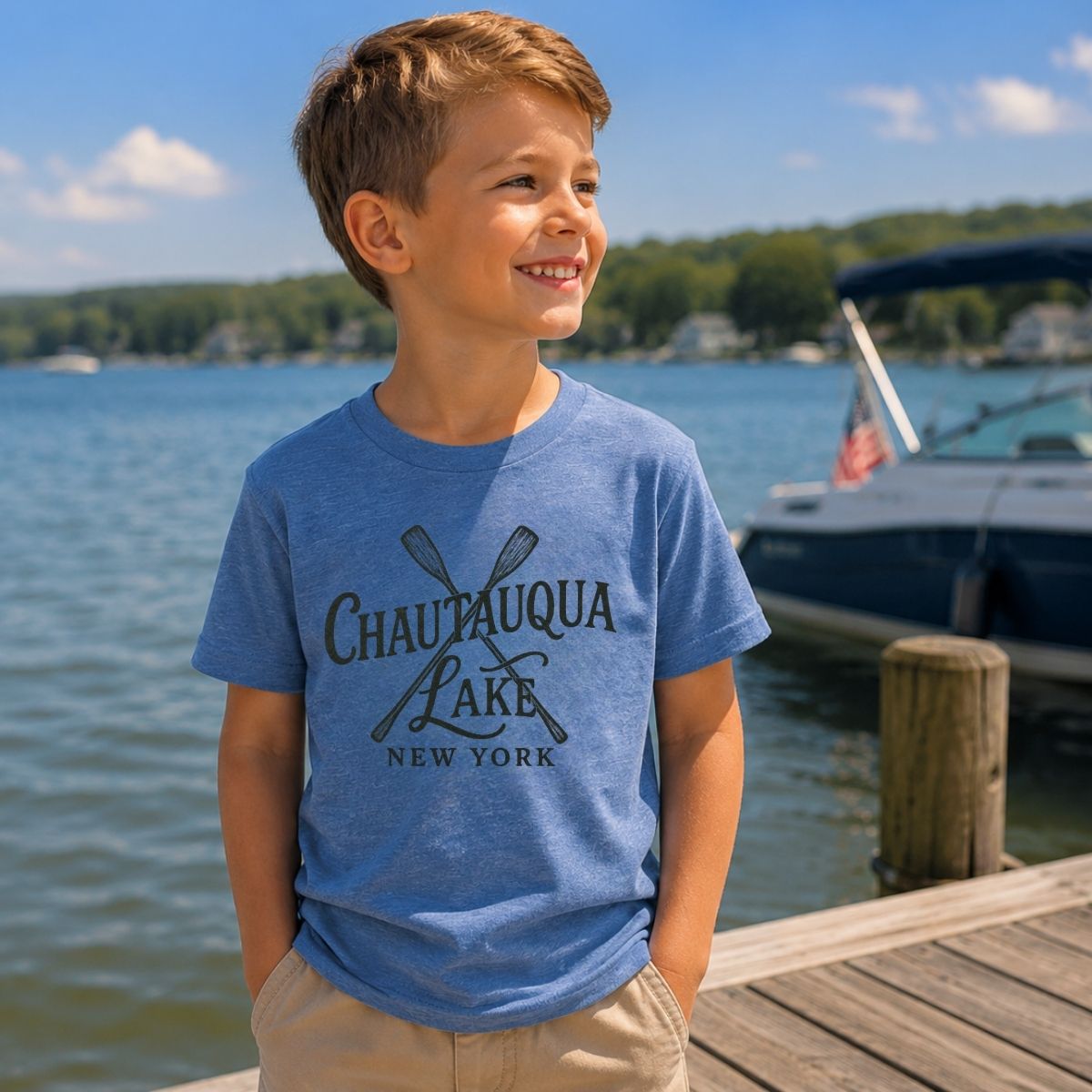 Child wearing a blue t-shirt with 'Chautauqua Lake, New York' text and sketched paddles on front standing by a lake with a boat in the background.