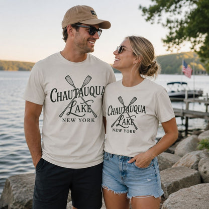Man and woman wearing beige "Chautauqua Lake New York" t-shirts with sketched paddles on front standing in front of a lake and dock.