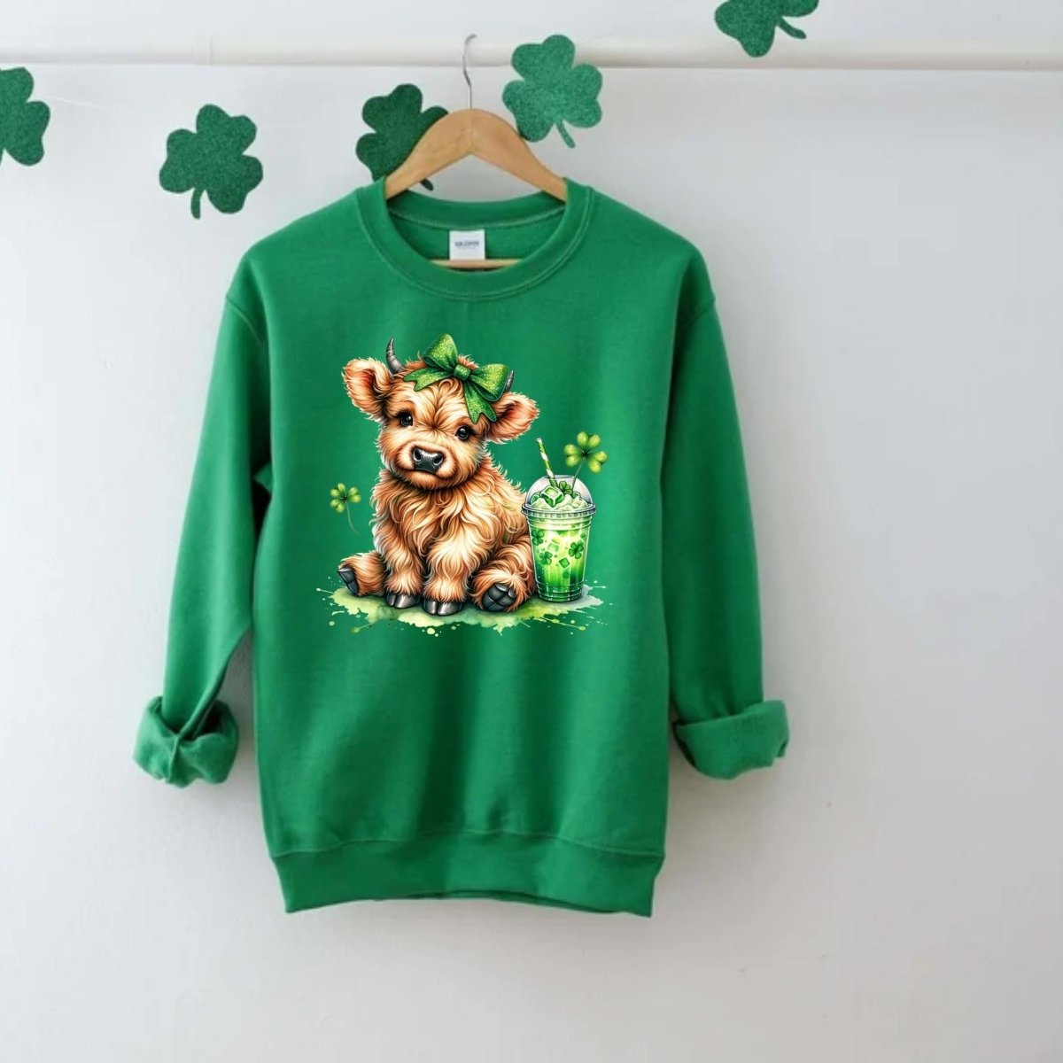 Irish Green crewneck sweatshirt with image of little brown cow in green hairbow with a shamrock drink and clovers on front hanging on a light gray background with clovers. 