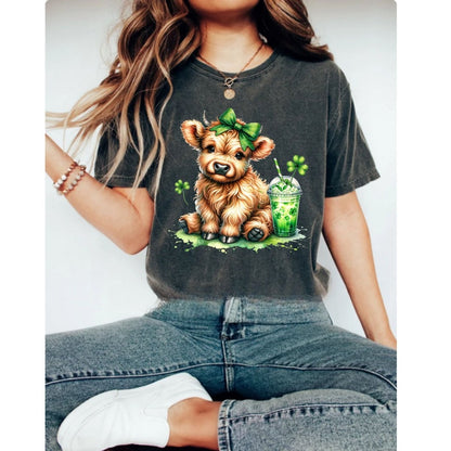 Girl wearing gray tshirt with image of brown cow in green hairbow with shamrock drink and clovers on front sitting on a gray background.