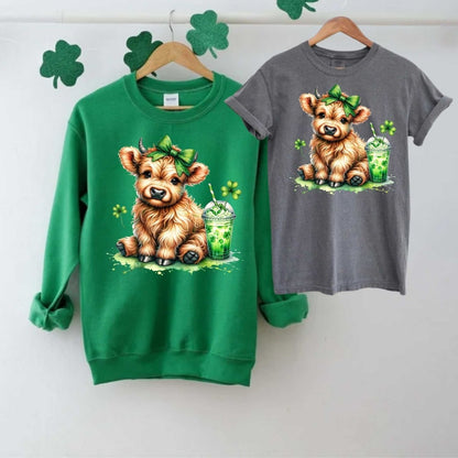 Green sweatshirt and gray tshirt with a cow in green bow with shamrock drink and clovers image on fronts on a light gray background. Little Gift Nook