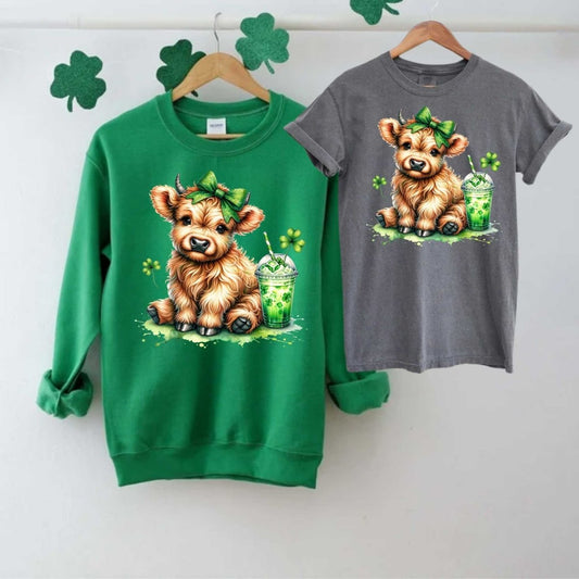 Green sweatshirt and gray tshirt with a cow in green bow with shamrock drink and clovers image on fronts on a light gray background. Little Gift Nook