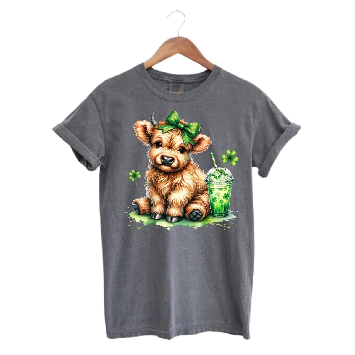 Gray tshirt with image of little brown cow in green hairbow with a shamrock drink and clovers on front hanging on a white background from hanger. 