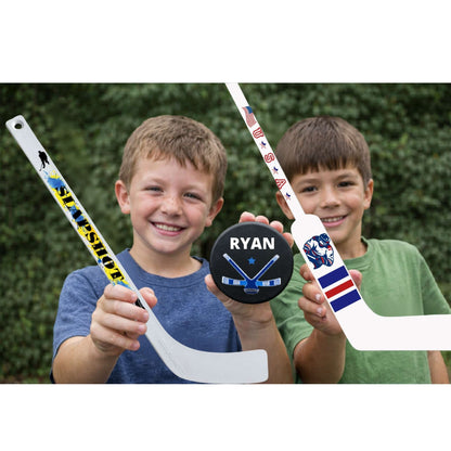 Two children holding mini hockey sticks and a puck with 'Ryan' on it against a green hedge background.