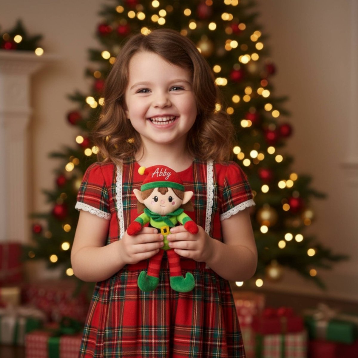 Little girl in plaid dress holding personalized name plush elf doll in front of Christmas tree background. 