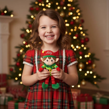 Little girl in plaid dress holding personalized name plush elf doll in front of Christmas tree background. 