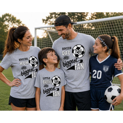 Sample custom t-shirts showing A family of four posing together on a soccer field wearing matching gray t-shirts with black and white soccer graphics and text. The shirts read 'SOCCER MOM AND biggest FAN', 'SOCCER DAD AND biggest FAN', and 'LITTLE BROTHER AND biggest FAN'.