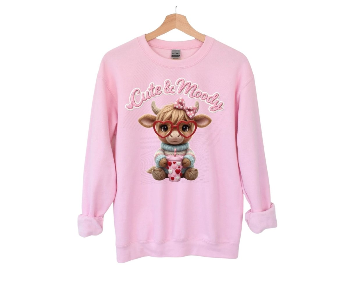 Pink sweatshirt with  "cute & moody" crochet style flat graphic cow in heart glasses on front hanging on white background.