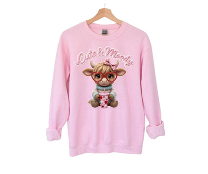 Pink sweatshirt with  "cute & moody" crochet style flat graphic cow in heart glasses on front hanging on white background.