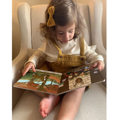Child reading a board book "Deer Camp" with colorful illustrations in a cozy setting.