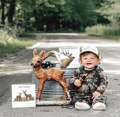 Baby dressed in camo sitting with deer figurine and Deer Camp board book on dirt road. 