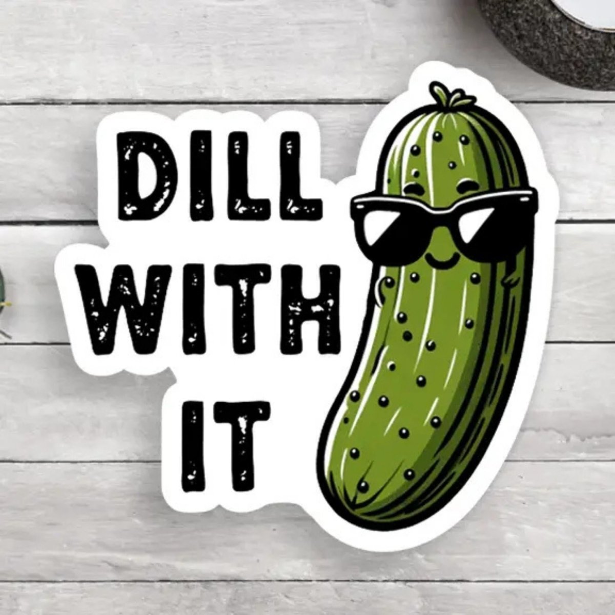 Green Pickle "Dill with it" sticker with white background sitting on a wood background.