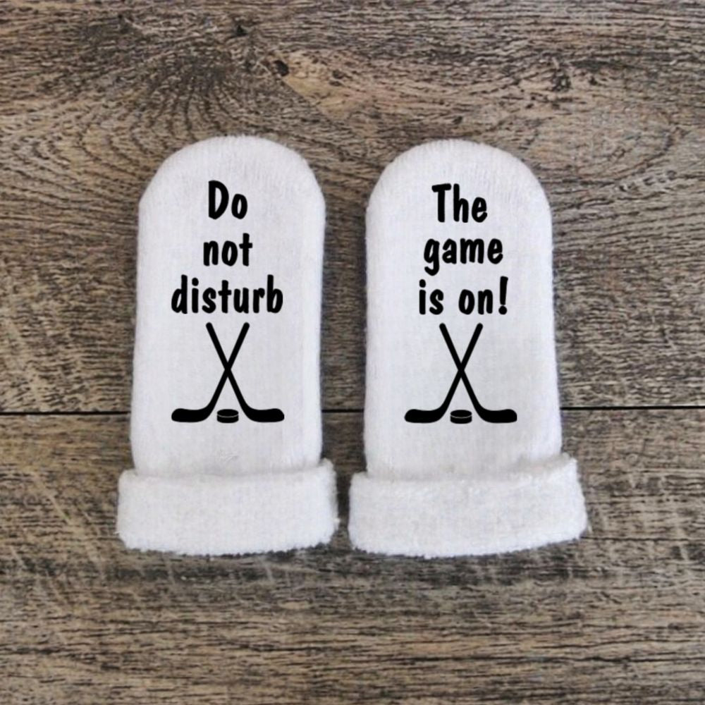 white baby socks with "Do not disturb the game is on" in black text and hockey graphic laying flat on wood background.