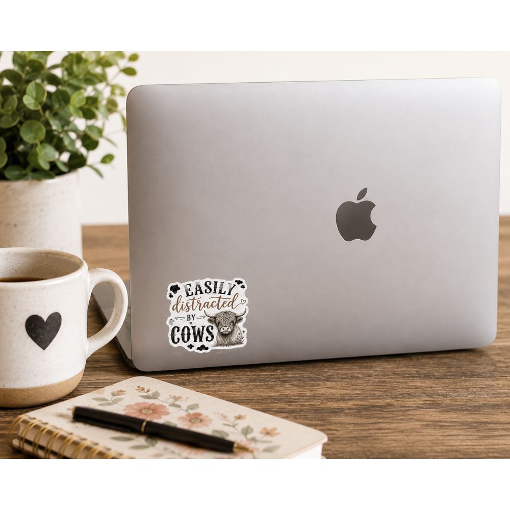 Laptop with a sticker on front of high land cow sketch and "easily distracted by cows in black and brown text. Sitting on a wooden surface with notebook and plant.