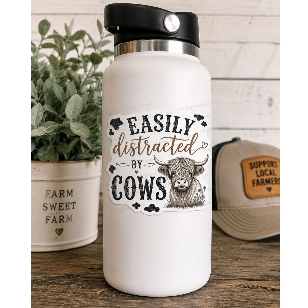 White water bottle with sticker on front of highland cow sketch and text "Easily distracted by cows" on a wooden surface with hat and plant.