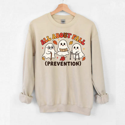 A beige crewneck sweatshirt hanging on a wooden hanger against a white wall. The sweatshirt features a graphic of three cute cartoon ghosts with autumn leaves and cane, walker and the text 'ALL ABOUT FALL (PREVENTION)' in a playful multicolor font.