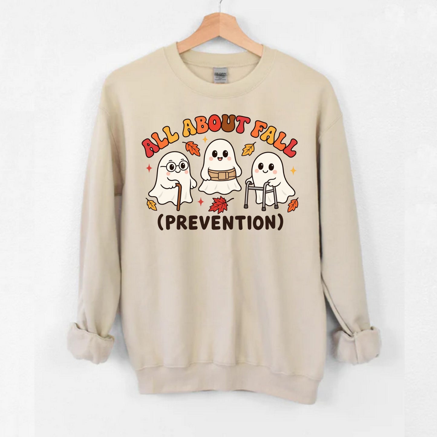A beige crewneck sweatshirt hanging on a wooden hanger against a white wall. The sweatshirt features a graphic of three cute cartoon ghosts with autumn leaves and cane, walker and the text 'ALL ABOUT FALL (PREVENTION)' in a playful multicolor font.