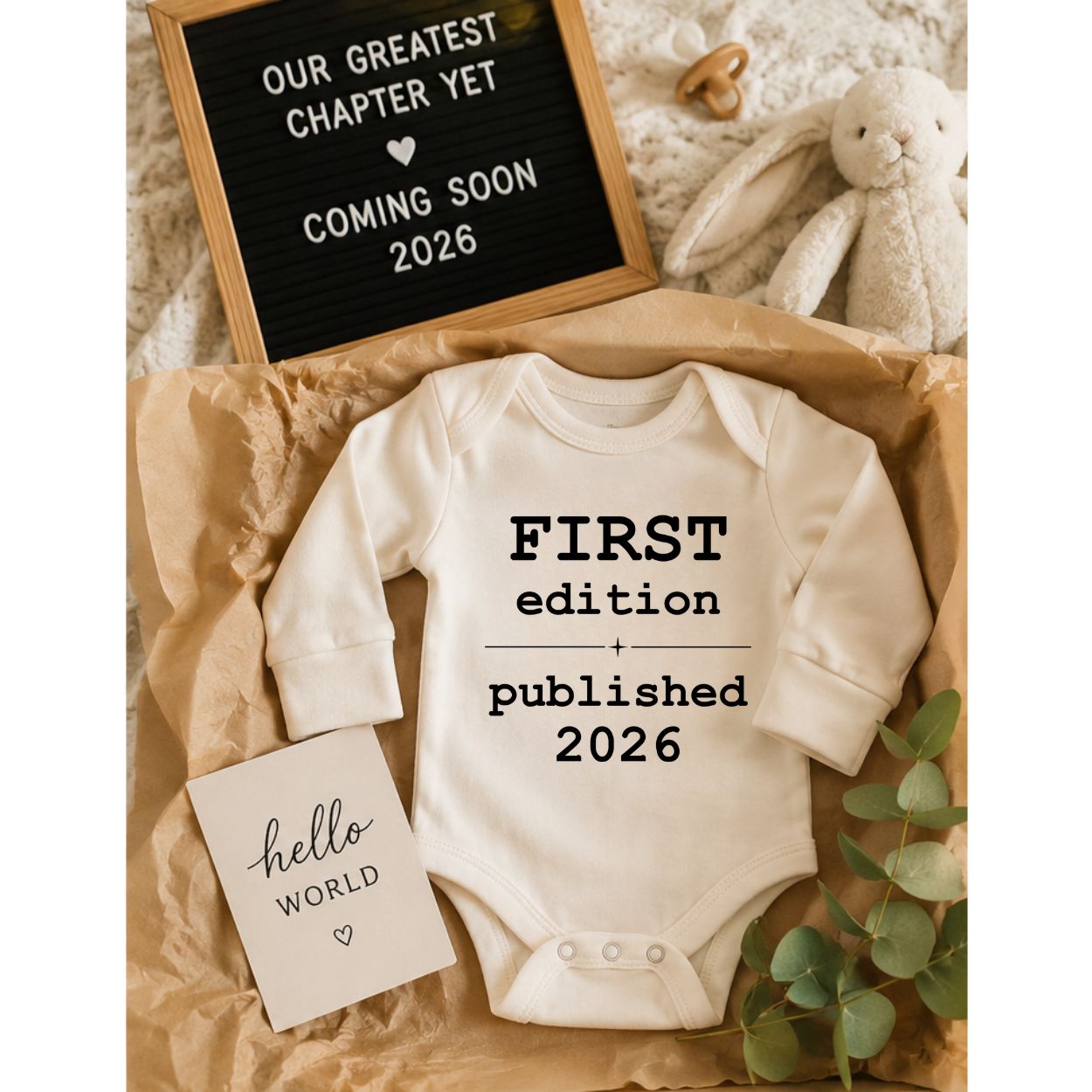 A cream-colored long-sleeve baby bodysuit with black text reading 'FIRST edition + published 2026' is displayed in a styled flat-lay arrangement on brown paper. The scene includes a letter board that says 'OUR GREATEST CHAPTER YET COMING SOON 2026', a small card reading 'hello WORLD', a plush bunny, and green eucalyptus leaves.