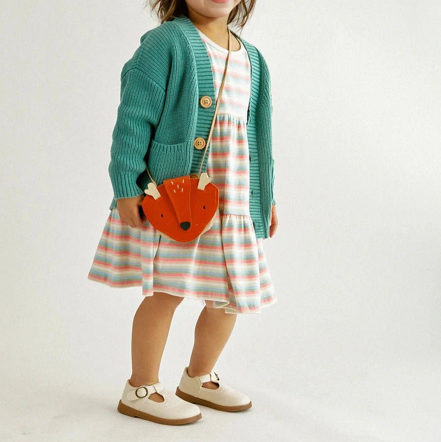 Little girl in striped dress and teal cardigan carrying orange little fox purse with strap standing on gray background.