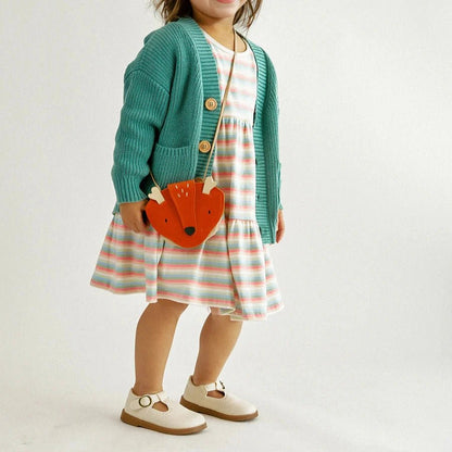 Little girl in striped dress and teal cardigan carrying orange little fox purse with strap standing on gray background.