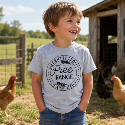 A young boy smiling outdoors on a farm, wearing a light gray heather t-shirt with a black circular graphic that reads 'CERTIFIED Free RANGE FARM KID.' Chickens are visible in the background near a wooden barn.