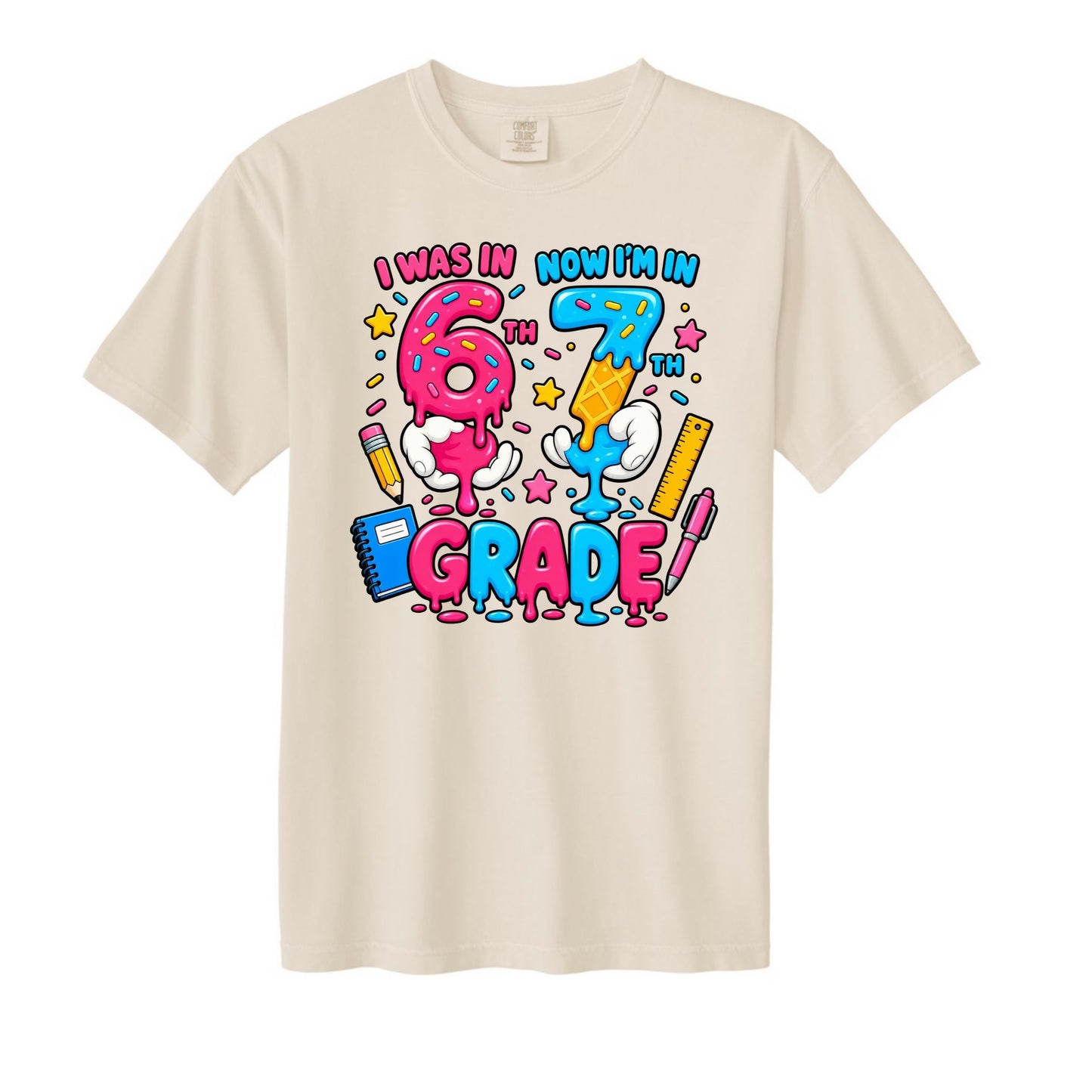 A cream-colored short-sleeve t-shirt with a colorful ice cream drip graphic on the front that reads 'I WAS IN NOW I'M IN GRADE' with cartoon hands and school supplies.