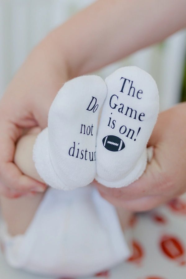 Close up of these white baby socks that say "Do Not Disturb the Game Is On" on bottoms while mom is holding legs up.