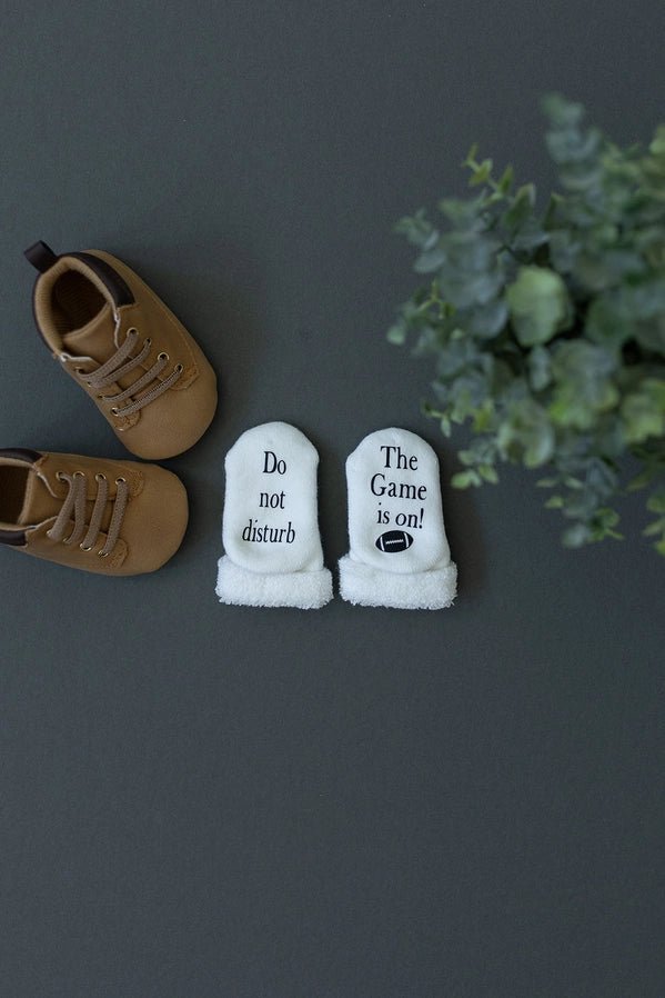 white baby socks that say "Do not disturb The Game is on" and little football image laying on a gray background with baby shoes.