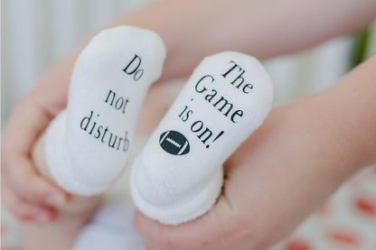 white baby socks with "Do not disturb" "The Game is on" written on bottoms on baby's feet behind held up.