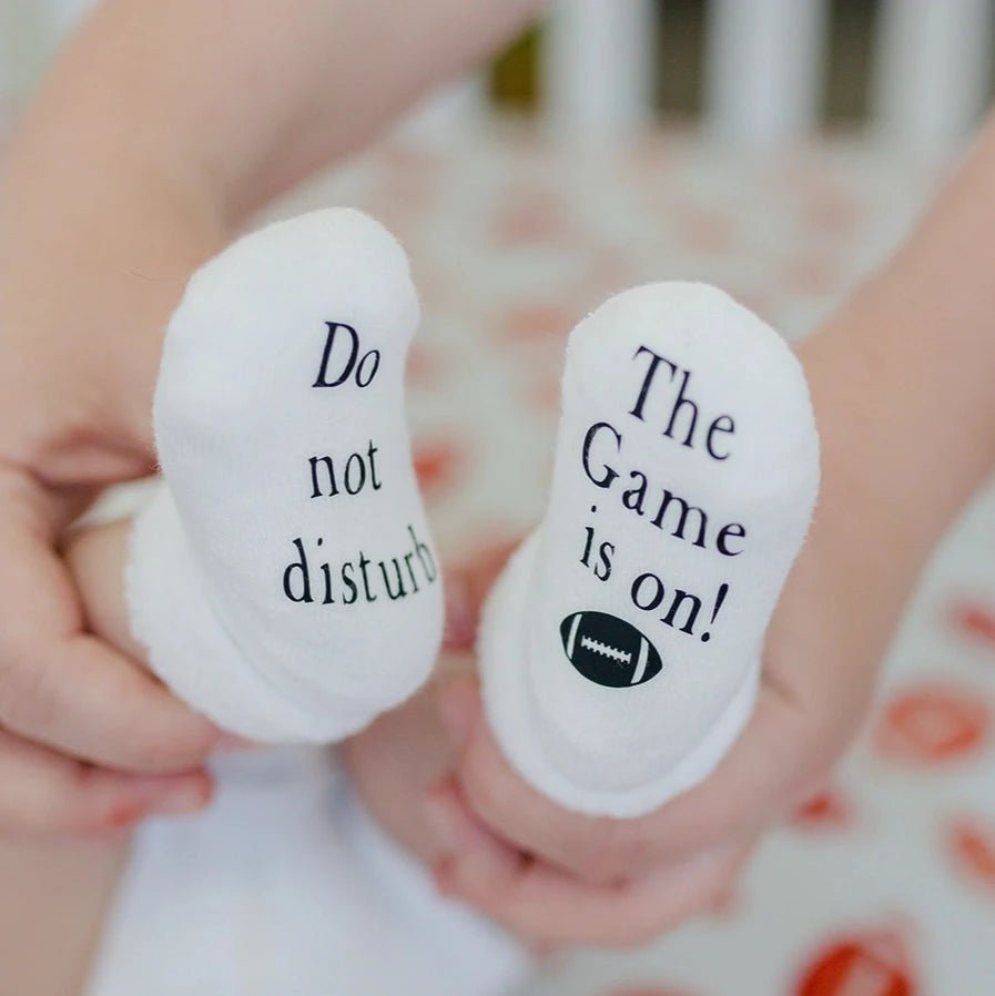 close up of baby feet wearing white socks with black text " Do not disturb The Game is on!" and little football on bottoms of them.