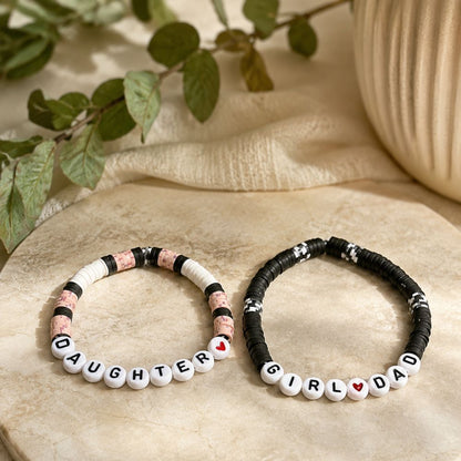 Two beaded bracelets with lettered beads "GIRL DAD" and "DAUGHTER" on a textured surface with greenery in the background.