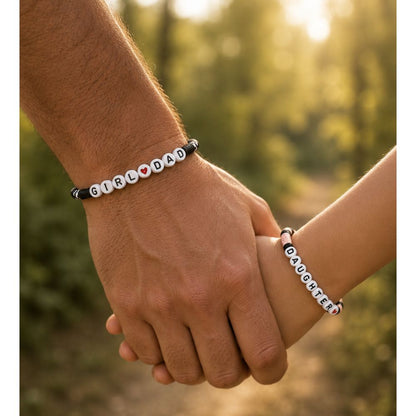 Two hands holding each other with 'GIRL DAD' and 'DAUGHTER' bracelets on their wrists in front of a greenery background.