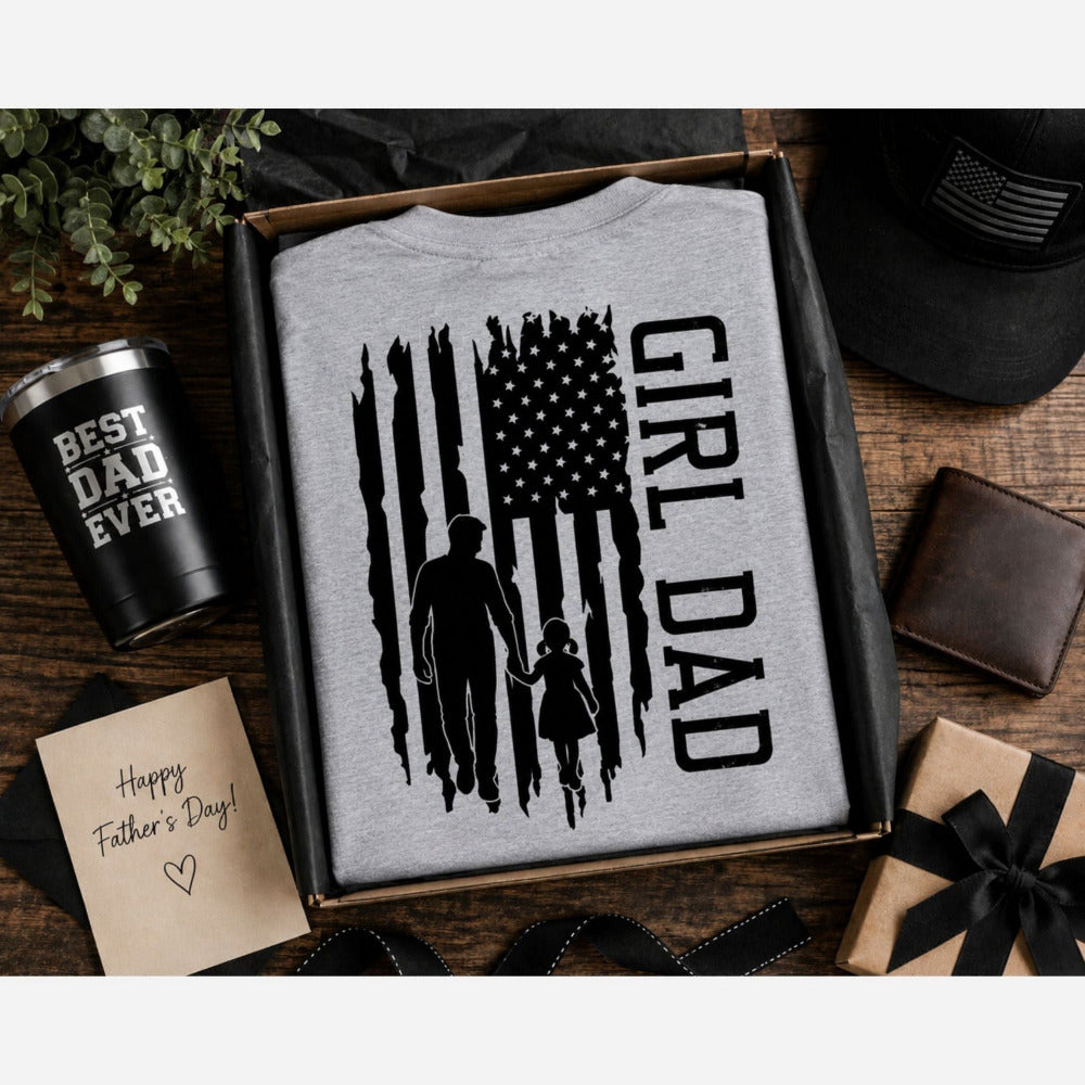 Gray t-shirt with 'Girl Dad' American flag design on back, folded in a box with Best dad ever tumbler and card on a wood surface.