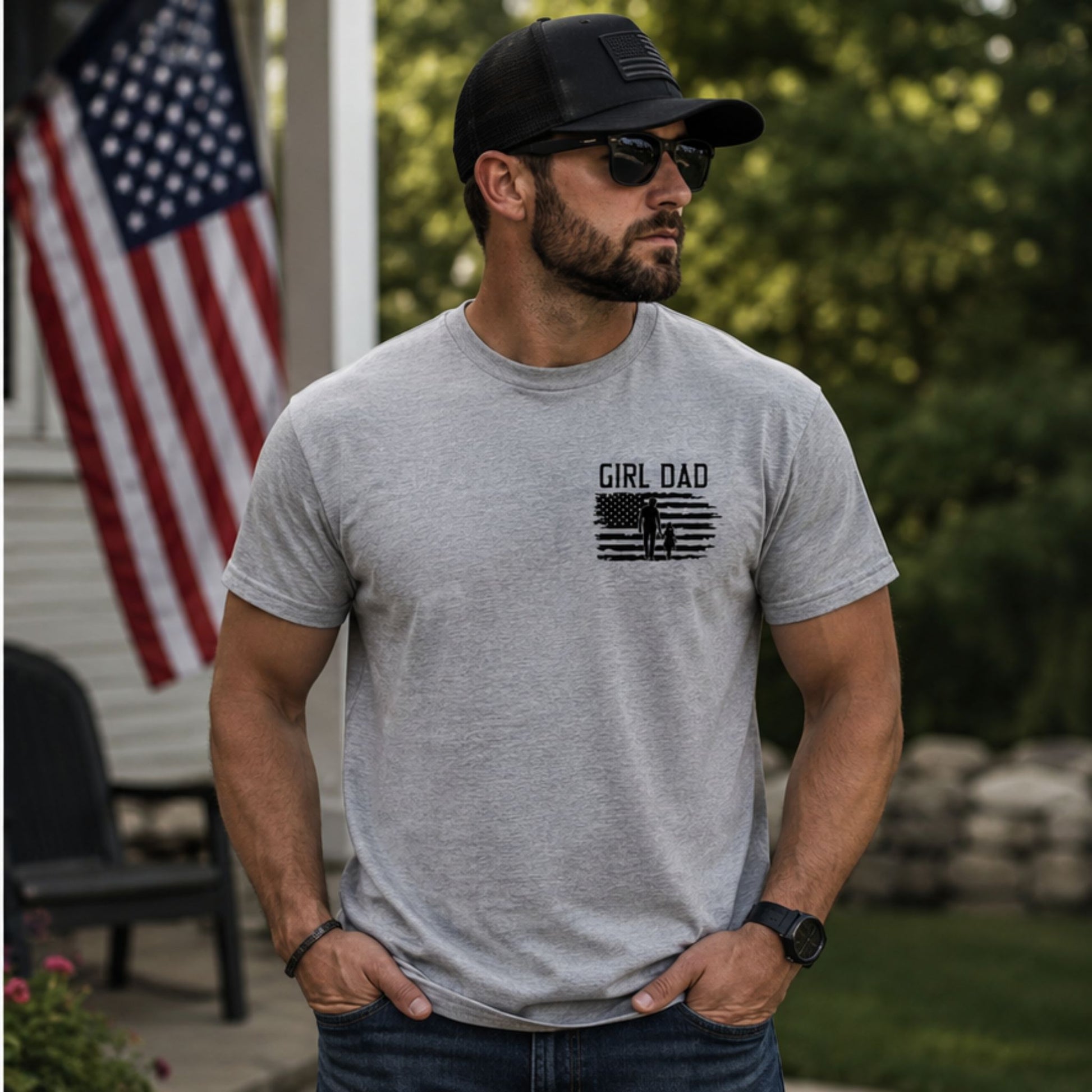 Man wearing heather gray t shirt with black silhouette graphic of American Flag and father & daughter and text "GIRL DAD" on front chest outside with a flag in the background.