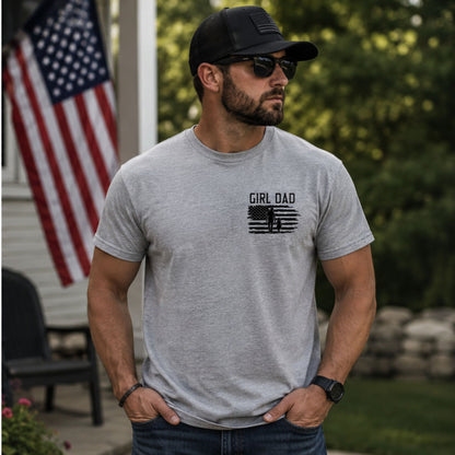 Man wearing heather gray t shirt with black silhouette graphic of American Flag and father & daughter and text "GIRL DAD" on front chest outside with a flag in the background.