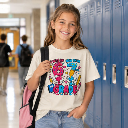 A young girl smiling in a school hallway next to blue lockers, wearing a cream-colored graphic t-shirt that reads 'I WAS IN 6TH NOW I'M IN 7TH GRADE' with colorful cartoon 67 hands ice cream drip theme, and carrying a pink backpack.