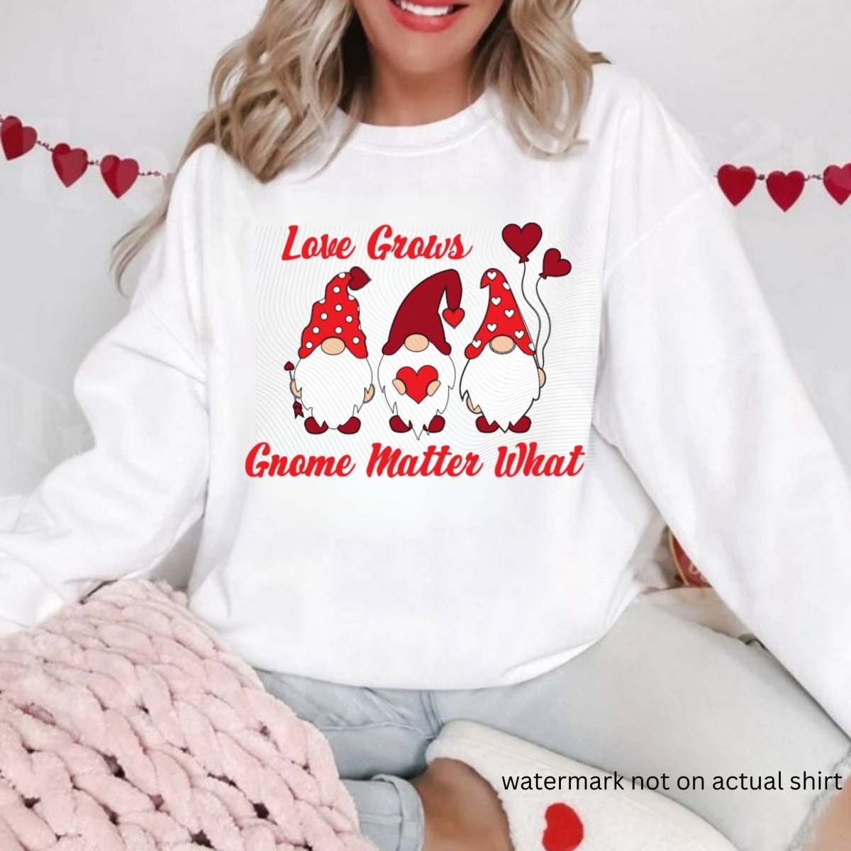 woman wearing white sweatshirt with 3 red gnomes and hearts "Love grows gnome matter what" on front sitting on white red heart background. Little Gift Nook