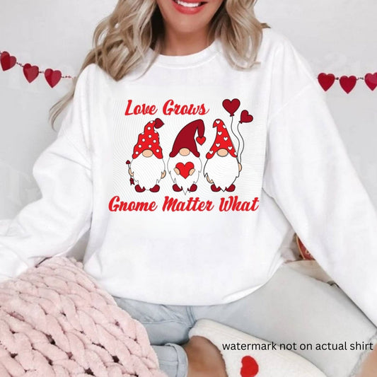 woman wearing white sweatshirt with 3 red gnomes and hearts "Love grows gnome matter what" on front sitting on white red heart background. Little Gift Nook