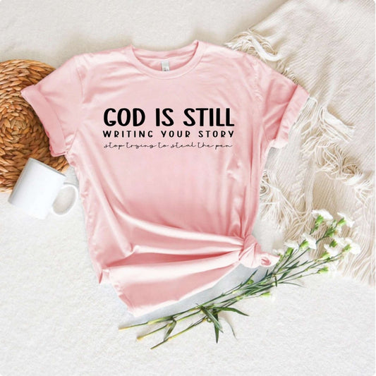 light pink tshirt with 'God is still writing your story. Stop trying to steal the pen" written in black text on front on white background with flowers. - Little Gift Nook