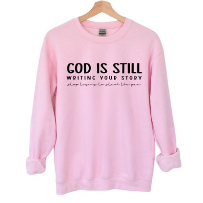 God Is Still Writing Your Story women's Pink Crewneck Sweatshirt. Light pink with black lettering on front. 