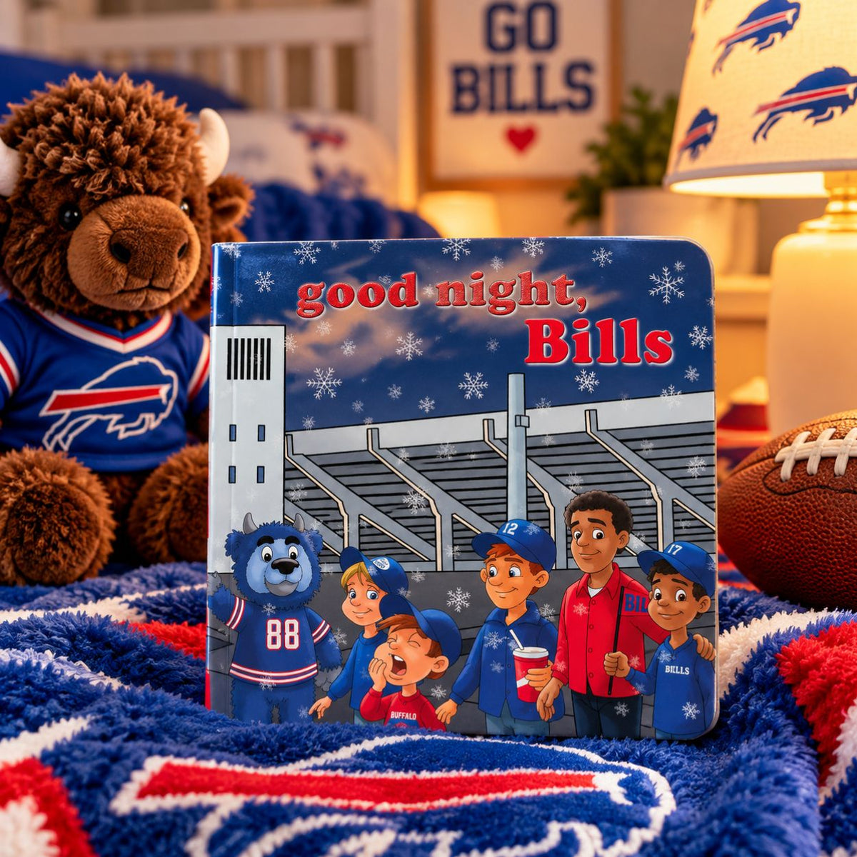 A children's board book titled 'good night, Bills' is displayed on a blue and red buffalo-themed blanket in a cozy bedroom setting. The book features cartoon characters and a stadium scene, surrounded by a plush buffalo toy, a football, and a lamp with Buffalo Bills branding.