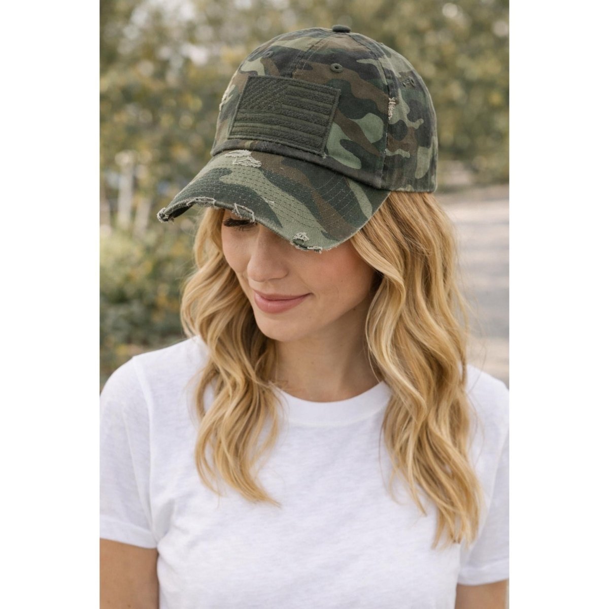 Woman wearing a green camo baseball cap with an American flag patch on front standing outdoors.