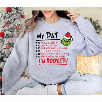 Woman wearing heather gray grinch daily schedule sweatshirt in front of a Christmas Holiday background. 