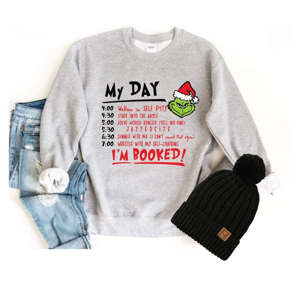 Gray Christmas Grinch Daily Schedule Sweatshirt with red and black text. Sitting on white background with jeans and pom hat.  
