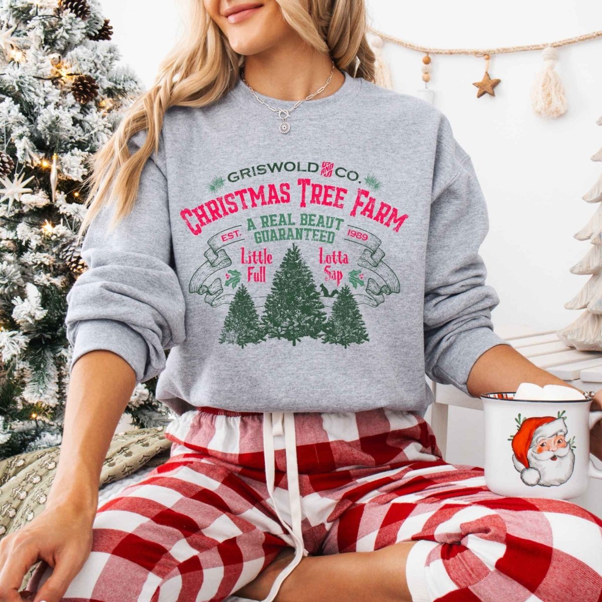 Woman wearing heather gray Griswold Christmas Tree Farm Sweatshirt sitting in front of Christmas tree background. 