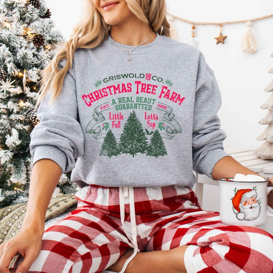 Woman wearing heather gray Griswold Christmas Tree Farm Sweatshirt sitting in front of Christmas tree background. 