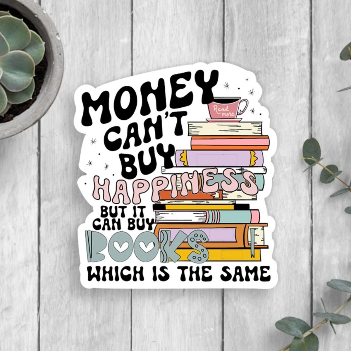 Sticker with books and "Money can't buy happiness but it can buy Books which is the same" quote on front laying flat on gray wood background.