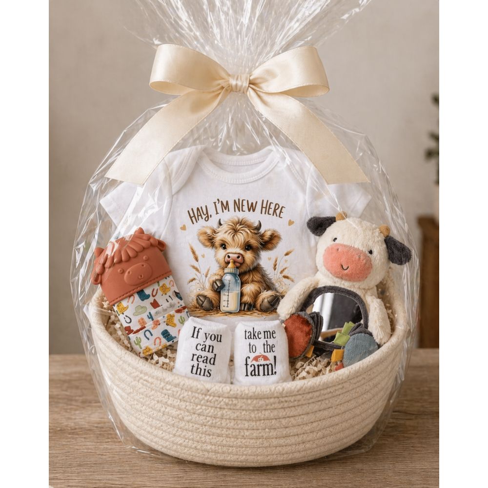 Baby gift basket with a cow onesie, cow mirror plush toy, cow teething mitt, and farm socks in clear cellophane wrap and rope basket on a wooden table top.